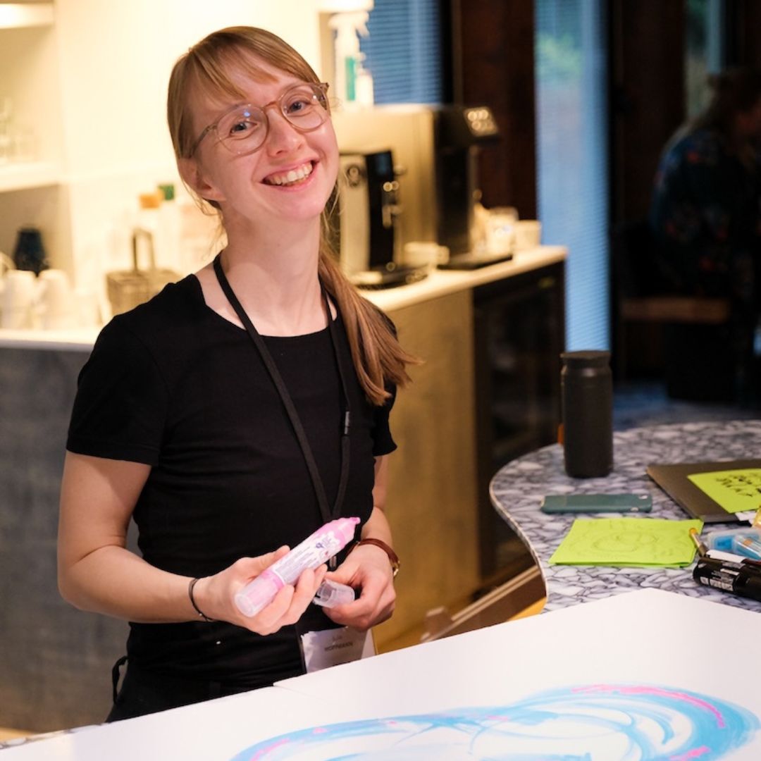 Julia Hoffmann lächelt in die Kamera, sie hat einen Stift in der Hand. Vor ihr ist ein Blatt Papier auf dem sie gemalt hat. 